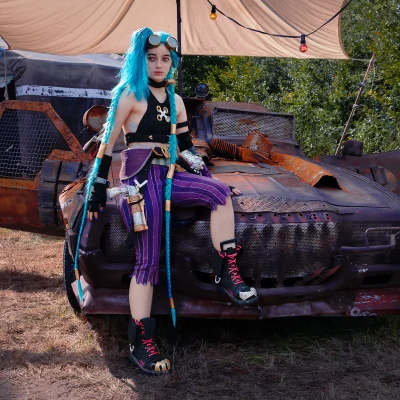 Fotoshooting: Jinx mit blauen Zöpfen, schwarzem Oberteil und lila-weiß gestreifter Hose sitzt auf einem rostigen Auto vor einem Zelt.