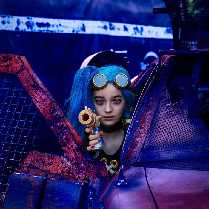 Jinx zielt mit einer Spielzeugwaffe durch die offene Tür eines rostigen, beschädigten Autos.