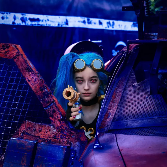Jinx zielt mit einer Spielzeugwaffe durch die offene Tür eines rostigen, beschädigten Autos.