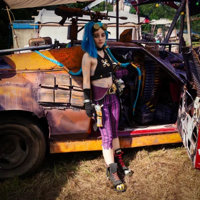 Cosplayerin mit blauen Zöpfen und violett gestreifter Hose lehnt an einem verrosteten, umgebauten Endzeit-Auto mit Panzerplatten