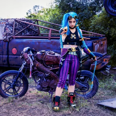 Jinx in punkigem Outfit mit blauen Zöpfen und Schutzbrille vor einem Motorrad richtet eine Pistole nach vorne.