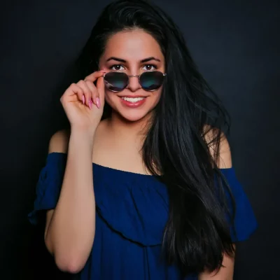 Frau mit langem dunklem Haar hebt eine Sonnenbrille leicht an und lächelt, trägt blaues schulterfreies Oberteil vor schwarzem Hintergrund.