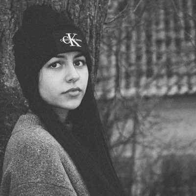 Schwarz-weiß Portrait einer Frau mit langen dunklen Haaren, die eine Mütze mit Calvin-Klein-Jeans-Logo trägt und seitlich an einen Baum lehnt.