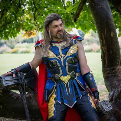 Mann in Thor-Kostüm mit rotem Umhang lehnt an einem Baum und hält einen Hammer; Fantasy-Fotoshooting in Soest