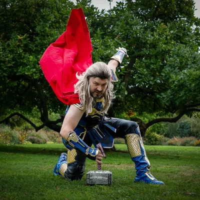 Thor mit rotem Umhang kniet auf Gras und hält Hammer auf dem Boden; Chris als kyurocosplay im Rosengarten in Soest