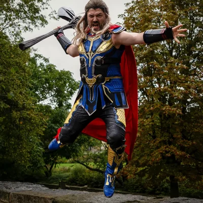 Chris als Thor mit rotem Umhang springt mit erhobenem Hammer über eine Steinmauer vor herbstlichen Bäumen