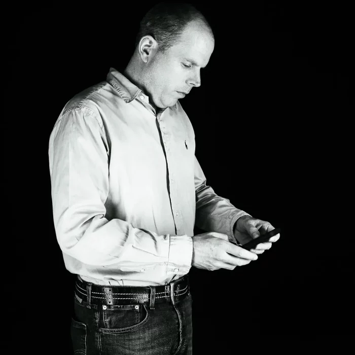 Business Portrait von Christian: Er steht seitlich, trägt Hemd und hält ein Smartphone mit beiden Händen, in Schwarz-Weiß.