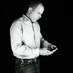 Business Portrait von Christian: Er steht seitlich, trägt Hemd und hält ein Smartphone mit beiden Händen, in Schwarz-Weiß.