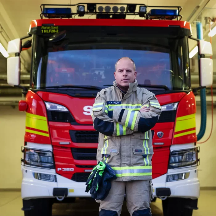 Mann in Feuerwehrschutzkleidung mit reflektierenden Streifen steht mit verschränkten Armen vor einem roten Feuerwehrauto im Depot bei einem Feuerwehr-Fotoshooting.