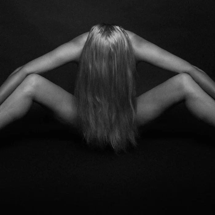 Shibari: Weibliches Model mit langen Haaren sitzt spreizend auf dem Boden, Handgelenke mit Bändern gefesselt, Kopf nach vorn geneigt.