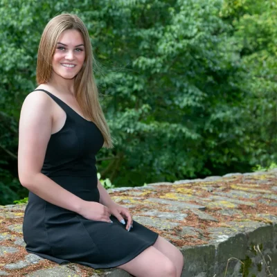 Weibliches Model in einem schwarzen ärmellosen Kleid sitzt lächelnd auf der moosbedeckten Stadtmauer von Soest vor grünem Laub.