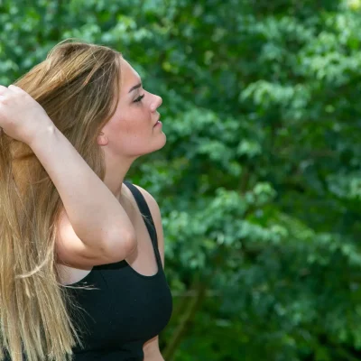 Weibliches Model mit langem blondem Haar, das ihr Haar mit der linken Hand nach hinten hält, seitliche Porträtaufnahme vor grünem Laub