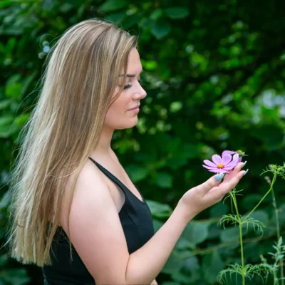 Weibliches Model mit langen blonden Haaren hält eine lila Blume und trägt ein schwarzes Top in einer grünen Außenaufnahme.