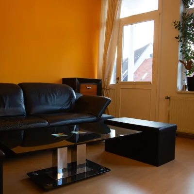 Ledersofa, Hocker und ein Glas-Couchtisch vor einer gelb gestrichenen Wand in einer Sitzecke mit Fenster und Pflanzen im Fotostudio