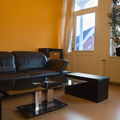 Ledersofa, Hocker und ein Glas-Couchtisch vor einer gelb gestrichenen Wand in einer Sitzecke mit Fenster und Pflanzen im Fotostudio