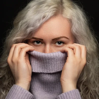 Weibliches Model mit lockigem, hellblondem Haar, das den hochgezogenen Kragen eines Strickpullovers vor dem Gesicht hält