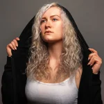 Franziska hebt die Kapuze eines schwarzen Kapuzenpullis an, trägt ein weißes ärmelloses Oberteil und blickt nach oben.