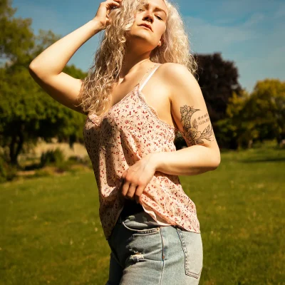 Frau mit lockigen blonden Haaren und Tattoo am Oberarm posiert im Freien in Sommerbekleidung aus Spitzen-Top und Jeans-Shorts.