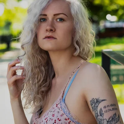 Portrait einer jungen Frau mit langen lockigen Haaren und floralem Tattoo auf dem Oberarm in einem sommerlichen Spitzenoberteil