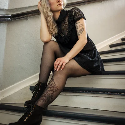 Franziska in schwarzem Kleid und schwarzer Strumpfhose sitzt auf einer Treppe mit sichtbaren Blumen-Tattoos am Bein und Arm.