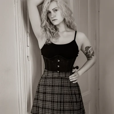 Weibliches Model mit lockigen Haaren in schwarzem Top, Korsage und kariertem Rock lehnt mit erhobenem Arm an einer Tür.