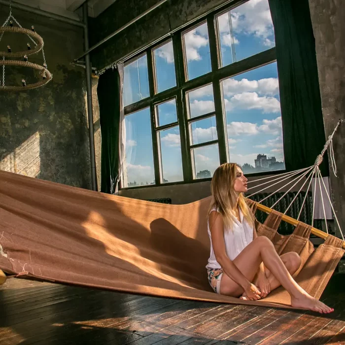 Junge Frau sitzt entspannt auf einer großen Hängematte in einem Loft in Minsk mit großen Fenstern und sichtbarem blauen Himmel.