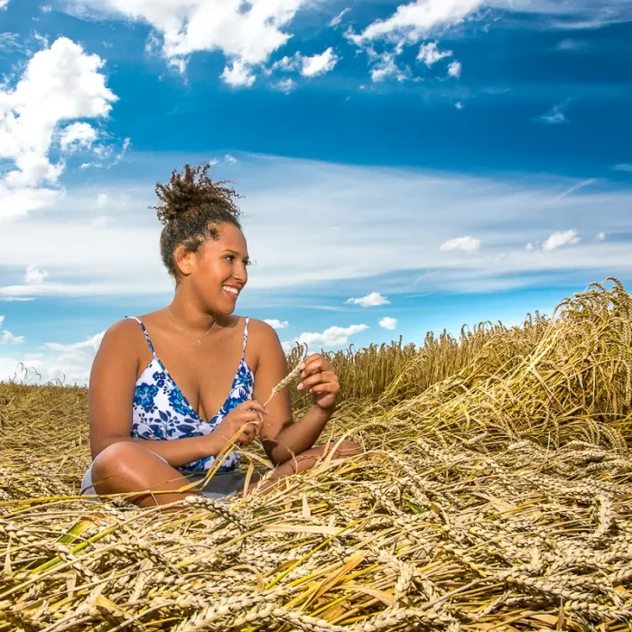 Weibliches Model mit blau-weißem Blumenkleid sitzt lächelnd im Kornfeld und hält eine Ähre in der Hand bei blauem Himmel.