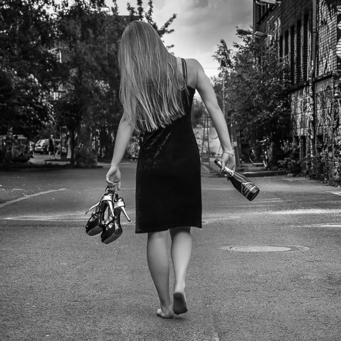 Jasmin von hinten fotografiert in schwarzem Kleid. Sie läuft barfuß eine Straße entlang, in der rechten Hand eine Sektflasche und in der linken ein Paar High Heels.