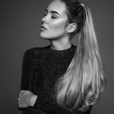 Porträtfotografie einer Frau mit langem Pferdeschwanz und geschlossenen Augen, verschränkten Armen vor dunkelgrauem Hintergrund