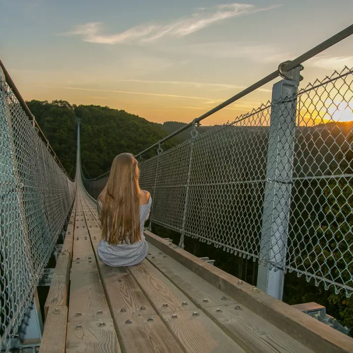 Frau mit langen Haaren sitzt bei Sonnenaufgang auf der Geierlay-Hängeseilbrücke und blickt zum Sonnenaufgang.