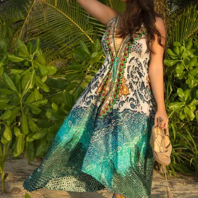 Weibliches Model in langem, gemustertem Kleid mit V-Ausschnitt hält ihre Sandalen in der rechten Hand und steht barfuß vor Palmen im Sand.