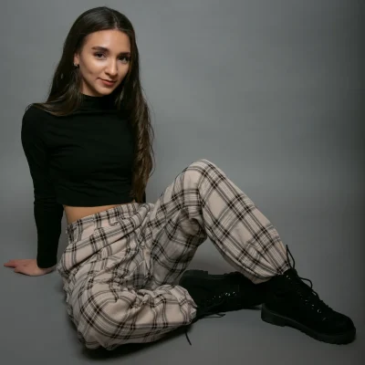 Weibliches Model sitzt auf grauem Boden, trägt karierte Hose, schwarzen Crop-Pullover und schwarze Stiefel, blickt leicht lächelnd zur Kamera.