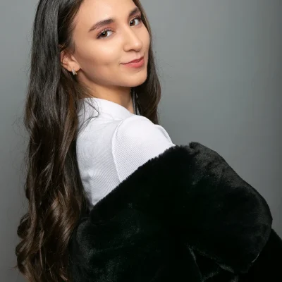 Weibliches Model mit langen, dunklen Haaren im Portrait, blickt über die Schulter, trägt weißen Pullover und schwarzen Pelzmantel.