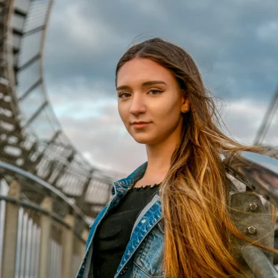 Porträt einer jungen Frau mit langem Haar und Jeansjacke vor der geschwungenen Konstruktion des Tiger and Turtle Denkmals