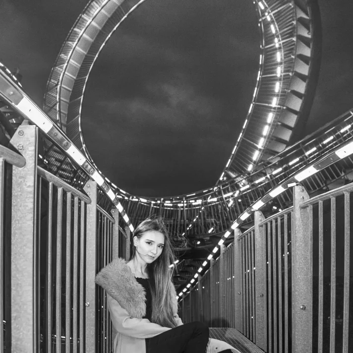 Magic Mountain: Gamze Kuva sitzt lässig auf der Treppeninstallation Tiger and Turtle, umgeben von Geländern und beleuchteten Schleifen, Schwarz-Weiß-Fotografie.