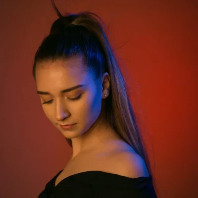 Porträt einer jungen Frau mit schwarzem schulterfreiem Oberteil, den Blick gesenkt, vor einem einfarbigen roten Hintergrund