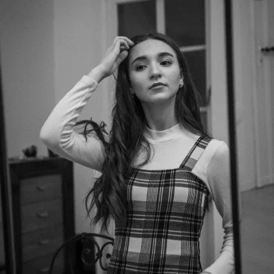 Model Gigi in kariertem Kleid und langem weißen Unterziehshirt betrachtet sich im Spiegel und berührt ihr Haar.