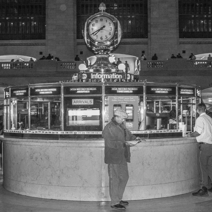 Schwarz-weißes Foto vom Informationsstand mit Uhr in der Grand Central Station in New York. Davor steht ein Mann mit Zeitschrift.