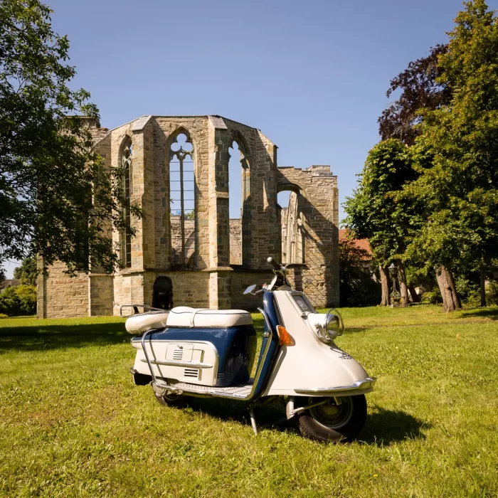 Heinkel-Roller aus den 1960er-Jahren im Vordergrund, stehend auf grüner Wiese vor Ruinenmauern mit gotischen Fenstern, umgeben von Bäumen