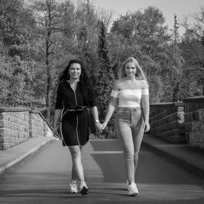 Zwei junge Frauen gehen in sommerlicher Kleidung Hand in Hand auf einer Steinbrücke über den Möhnesee, umgeben von Bäumen.