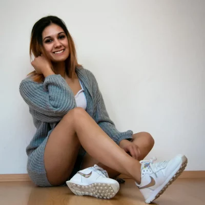 Weibliches Model in hellgrauem Strickpullover und Turnschuhen sitzt entspannt auf hellem Holzboden vor einer weißen Wand.