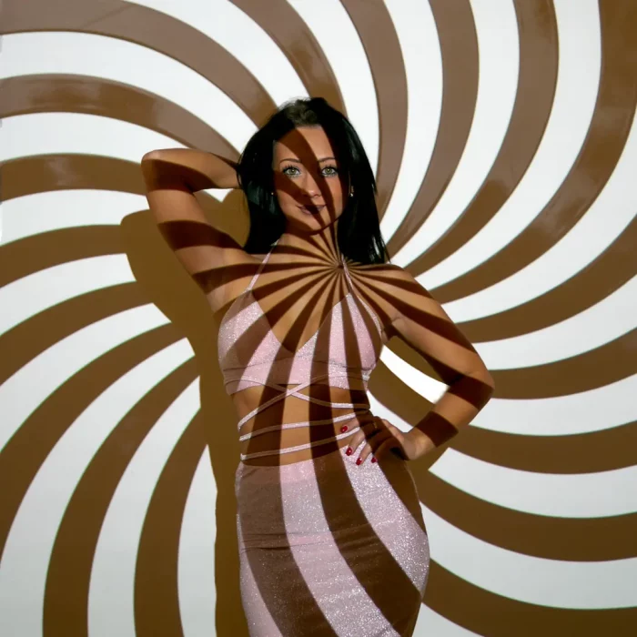Frau trägt ein Kleid mit Cut-Outs im Taillenbereich vor einem braun-weißen Spiralmuster, das sich über Körper und Hintergrund erstreckt.