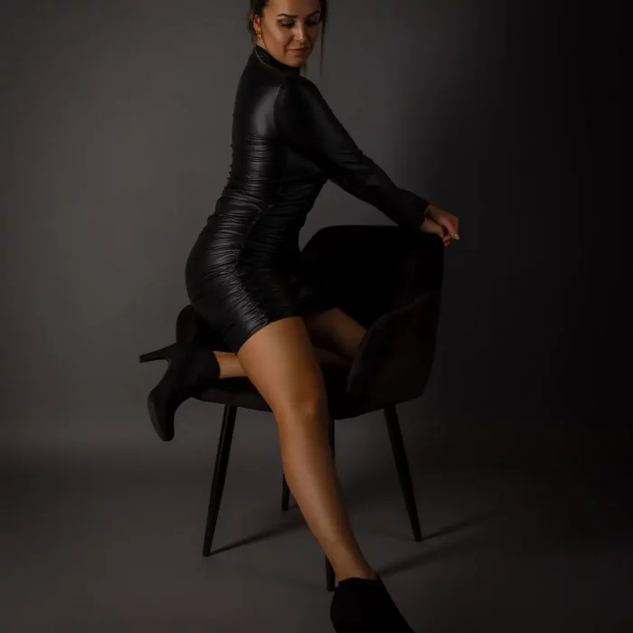 Portraitfotografie eines weiblichen Models im schwarzen Lederkleid, das kniend auf einem dunklen Stuhl posiert, vor neutralem Hintergrund