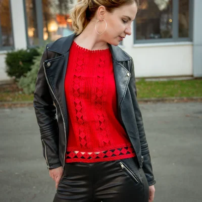 Junge Frau in rotem Strickoberteil und schwarzer Lederjacke blickt seitlich nach unten, draußen vor einem Gebäude.
