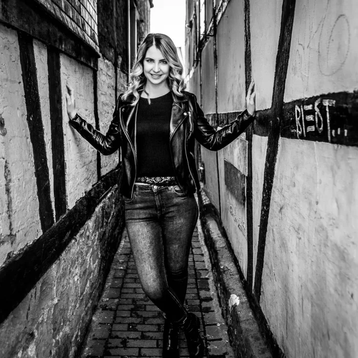 Portraitfotografie einer Frau in schwarzer Lederjacke und Jeans, stehend in einer schmalen Gasse, schwarz-weiß aufgenommen