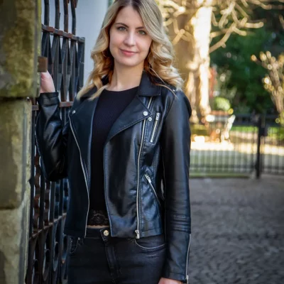 Frau in schwarzer Lederjacke und dunkler Hose lehnt an schmiedeeisernem Parktor, Hintergrund mit Bäumen und gepflastertem Weg