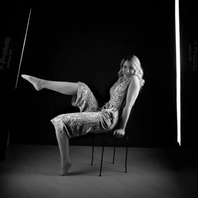 Frau in einem schwarz-weiß gestreiften Jumpsuit, sitzend auf einem Stuhl mit ausgestrecktem Bein vor dunklem Hintergrund