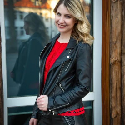 Porträtaufnahme von Jana Zebrowski mit rotem Oberteil und schwarzer Lederjacke, stehend vor einem Fenster mit Spiegelung