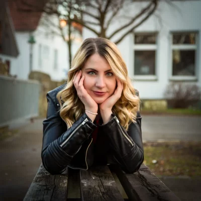 Junge Frau mit langen gewellten Haaren und schwarzer Lederjacke stützt das Kinn auf die Hände, Porträtfotografie im Freien auf einer Holzbank