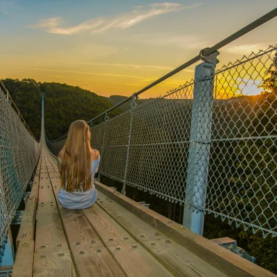 Frau mit langen Haaren sitzt bei Sonnenaufgang auf der Geierlay-Hängeseilbrücke und blickt zum Sonnenaufgang.